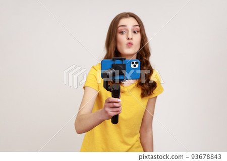Young attractive woman with camera phone on stabilizer shoots video for her vlog, making kissing gesture, wearing yellow casual style T-shirt. Indoor studio shot isolated on gray background. 93678843