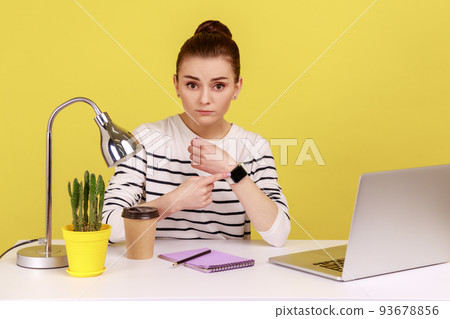 Serious woman employee sitting in office and pointing to watch on her hand, looking disgruntled, exhausted of overtime work and need break. Indoor studio studio shot isolated on yellow background. Serious woman employee sitting in office and pointing to watch on her hand, looking disgruntled, exhausted of overtime work and need break. Indoor studio studio shot isolated on yellow background. 93678856