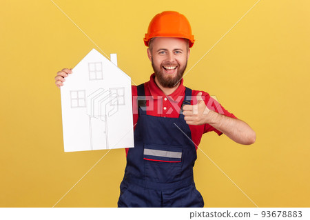 Portrait of builder man holding paper house, offering help in relocation, recommend home repair services, showing thumb up, wearing overalls and cap. Indoor studio shot isolated on yellow background. Portrait of builder man holding paper house, offering help in relocation, recommend home repair services, showing thumb up, wearing overalls and cap. Indoor studio shot isolated on yellow background. 93678883