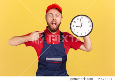 Shocked surprised man worker standing and pointing at big wall clock in his hands, looking at camera with open mouth, wearing overalls and red cap. Indoor studio shot isolated on yellow background. Shocked surprised man worker standing and pointing at big wall clock in his hands, looking at camera with open mouth, wearing overalls and red cap. Indoor studio shot isolated on yellow background. 93678895