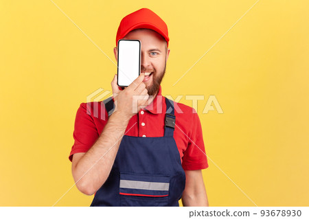 Portrait of happy smiling joyful bearded worker man standing and covering eye with cell phone with blank screen for promotional text. Indoor studio shot isolated on yellow background. 93678930