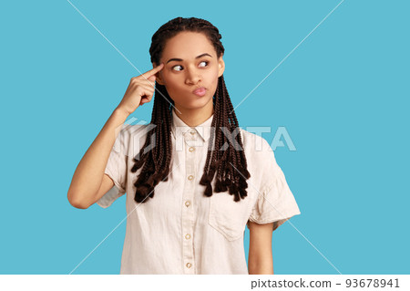 Positive woman with black dreadlocks holding finger near head temple and gesturing stupid idiot, looking displeased with crazy idea, wearing white shirt. Indoor studio shot isolated on blue background Positive woman with black dreadlocks holding finger near head temple and gesturing stupid idiot, looking displeased with crazy idea, wearing white shirt. Indoor studio shot isolated on blue background 93678941