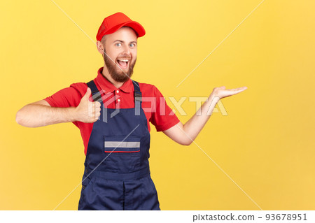 Portrait of optimistic handyman standing and presenting copy space for advertisement on his palm, showing thumb up, wearing overalls and red cap Indoor studio shot isolated on yellow background. 93678951