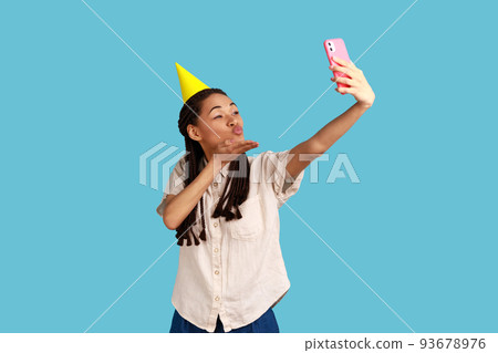 Romantic woman in cone broadcasting livestream, sending air kiss to followers, recording video for blog during celebration birthday, wearing white shirt. Indoor studio shot isolated on blue background Romantic woman in cone broadcasting livestream, sending air kiss to followers, recording video for blog during celebration birthday, wearing white shirt. Indoor studio shot isolated on blue background 93678976