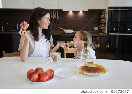 Crop shot of mother in red and her daughter having colorful donuts sitting at kitchen. Dieting concept and junk food. Crop shot of mother in red and her daughter having colorful donuts sitting at kitchen. Dieting concept and junk food. 93678977