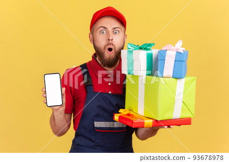 Portrait of shocked courier man wearing blue overalls holding stack of present boxes and smart phone with blank screen for advertisement. Indoor studio shot isolated on yellow background. Portrait of shocked courier man wearing blue overalls holding stack of present boxes and smart phone with blank screen for advertisement. Indoor studio shot isolated on yellow background. 93678978