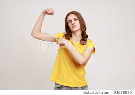 Portrait of proud strong and independent woman of young age in casual style clothing standing and pointing finger at her biceps, showing her power. Indoor studio shot isolated on gray background. Portrait of proud strong and independent woman of young age in casual style clothing standing and pointing finger at her biceps, showing her power. Indoor studio shot isolated on gray background. 93678980
