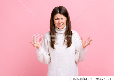 Portrait of angry female standing with raised arms, expressing aggression, screaming, frowning face, wearing white casual style sweater. Indoor studio shot isolated on pink background. 93678994