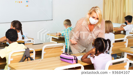 Teacher woman in protective mask helping schoolkid during lesson 93684107