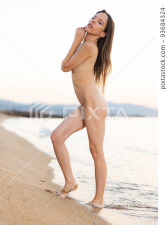 Fully naked brunette standing on sandy shore by ocean 93684324