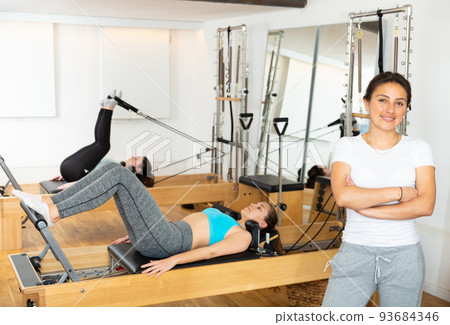 Female pilates trainer posing against the background of training people in gym Female pilates trainer posing against the background of training people in gym 93684346