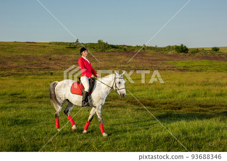 Young woman rider, wearing red redingote and white breeches, with her horse in evening sunset light. Young woman rider, wearing red redingote and white breeches, with her horse in evening sunset light. 93688346