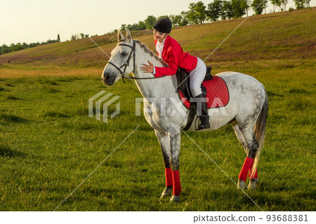 Young woman rider, wearing red redingote and white breeches, with her horse in evening sunset light. Young woman rider, wearing red redingote and white breeches, with her horse in evening sunset light. 93688381