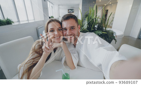 Point of view shot of attractive loving couple recording video while relaxing in day spa together. Happy young people are smiling, laughing and kissing looking at camera. 93689107