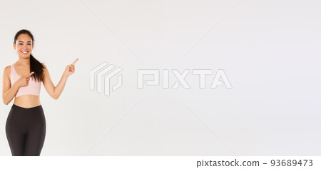 Full length of smiling, cute slim asian girl in sportswear, pointing fingers upper right corner, inviting to buy gym membership, showing workout offer, fitness coach show healthy diet recommendations 93689473