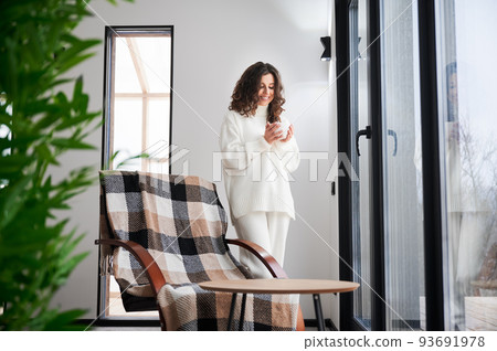 Curly young woman enjoying weekends inside contemporary barn house. Happy female tourist standing near chair, holding cup of tea in new cottage. 93691978