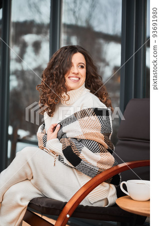 Curly young woman resting on terrace of modern barn house in the mountains. Happy female tourist in plaid sitting on chair, enjoying in new cottage in winter. Curly young woman resting on terrace of modern barn house in the mountains. Happy female tourist in plaid sitting on chair, enjoying in new cottage in winter. 93691980