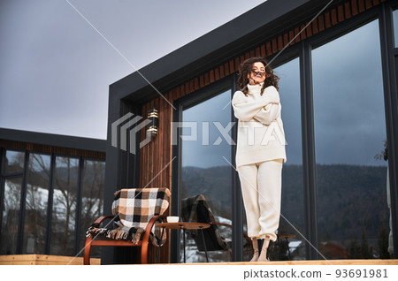 Curly young woman resting on terrace of modern barn house in the mountains. Happy female tourist enjoying in new cottage in winter. Curly young woman resting on terrace of modern barn house in the mountains. Happy female tourist enjoying in new cottage in winter. 93691981