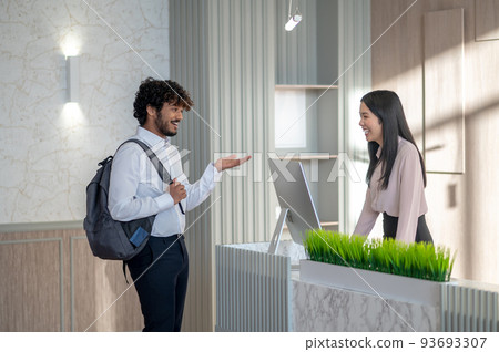 Indian young man with backpack talking to a girl on reception Indian young man with backpack talking to a girl on reception 93693307