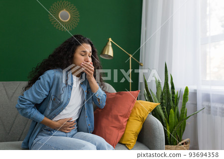 Young beautiful hispanic woman sick at home sitting on sofa with nausea and stomach ache sitting on sofa in living room near window with green wall Young beautiful hispanic woman sick at home sitting on sofa with nausea and stomach ache sitting on sofa in living room near window with green wall 93694358