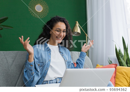 Young beautiful hispanic woman with curly hair at home using laptop for video call, woman smiling and chatting with friends remotely sitting on sofa in living room with green wall Young beautiful hispanic woman with curly hair at home using laptop for video call, woman smiling and chatting with friends remotely sitting on sofa in living room with green wall 93694362