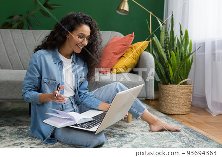 Young beautiful Hispanic female student studying remotely online at home, taking lecture notes sitting on floor in living room with green wall, woman taking courses studying online Young beautiful Hispanic female student studying remotely online at home, taking lecture notes sitting on floor in living room with green wall, woman taking courses studying online 93694363