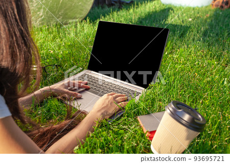 Close up hands on keyboard. Woman working on laptop pc computer with blank black empty screen to copy space in park on green grass sunshine lawn outdoors. Close up hands on keyboard. Woman working on laptop pc computer with blank black empty screen to copy space in park on green grass sunshine lawn outdoors. 93695721