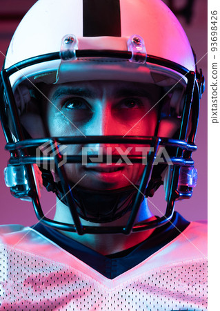 Close-up portrait of young man, american football player in helmet posing isolated over purple background neon light. 93698426