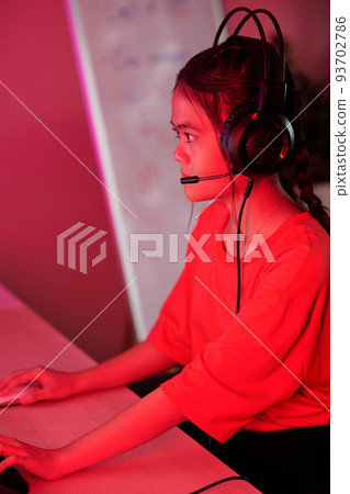 Preteen girl wearing heaphones with microphone when playing videogame on computer and streaming 93702786
