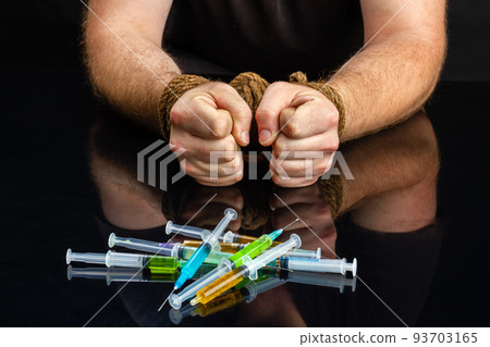 Drug addiction dependence and hostage concept. Bound male hands. Pile of syringes with various drugs. 93703165