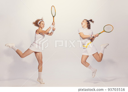 Portrait of two active young women in retro uniform playing badminton isolated over grey studio background 93703516