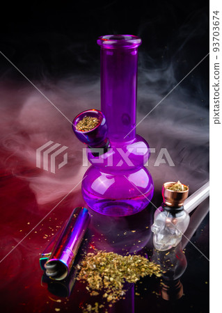 Vertical shot of glass bong with smoking pipe and pile of weed. Set of marijuana and smoking devices. Vertical shot of glass bong with smoking pipe and pile of weed. Set of marijuana and smoking devices. 93703674