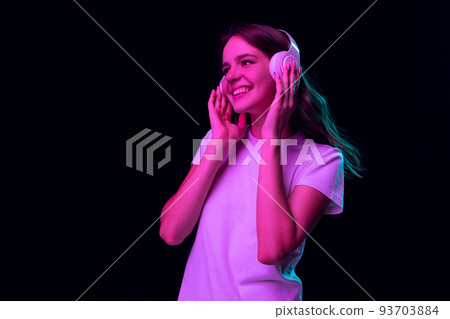 Music. Portrait of pretty young girl, student wearing white tee isolated on dark background in pink neon light. Concept of beauty, art, fashion, emotions Music. Portrait of pretty young girl, student wearing white tee isolated on dark background in pink neon light. Concept of beauty, art, fashion, emotions 93703884
