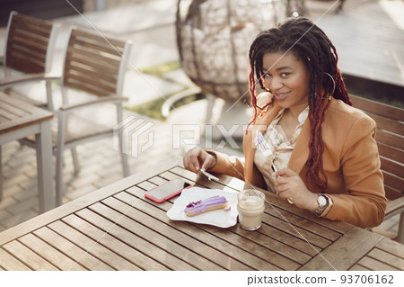 Smiling african american woman drinking coffee and eating dessert in outdoor cafe 93706162