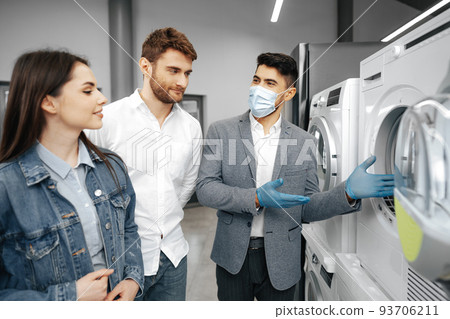 Salesman in hypermarket wearing medical mask demonstrates his clients a new washing machine Salesman in hypermarket wearing medical mask demonstrates his clients a new washing machine 93706211