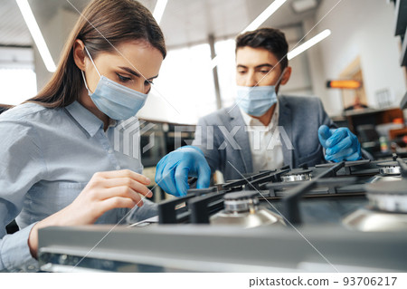 Male salesman in hypermarket describes features of gas stove to female client, both wearing face masks Male salesman in hypermarket describes features of gas stove to female client, both wearing face masks 93706217