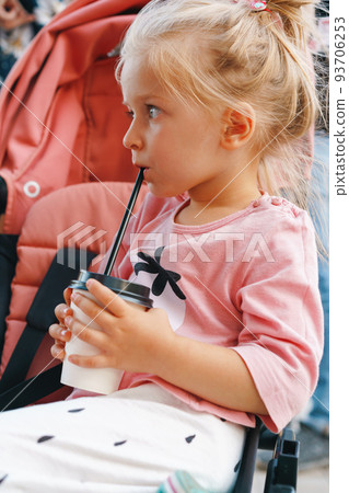 Blonde toddler girl sitting in the stroller and drinking from disposable cup 93706253