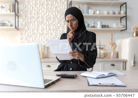Arab woman at home doing paperwork, concentrated Muslim woman in hijab holding bills in hands, calculating household budget paying loan, using laptop in kitchen at home 93706447