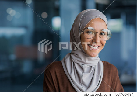 Close up photo portrait of beautiful young muslim woman, woman in hijab and glasses smiling and looking at camera, businesswoman working inside modern office building 93706484