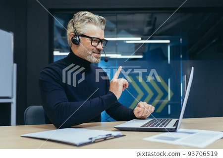 Senior and experienced gray-haired boss working in modern office, man with headset talking on video call using laptop while sitting at table, businessman in online meeting talking colleagues remotely 93706519