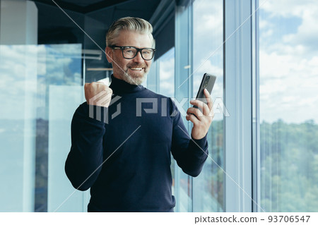 Portrait of successful senior experienced investor, man working in office, gray-haired businessman in glasses smiling and looking at camera, broker holding phone in hands, celebrating victory triumph 93706547