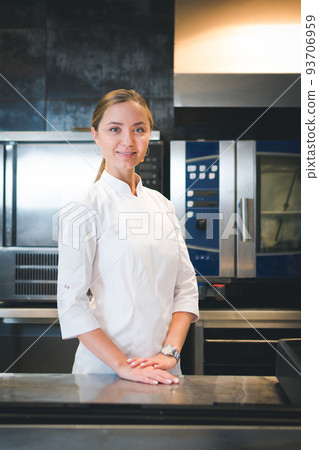 Portrait of confident and smiling young woman chef dressed in white uniform, professional kitchen are on background Portrait of confident and smiling young woman chef dressed in white uniform, professional kitchen are on background 93706959