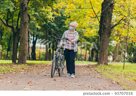 Senior beautiful blonde woman rides a bike in the park, in the woods, in nature in a medical mask during quarantine, leads a healthy active lifestyle Senior beautiful blonde woman rides a bike in the park, in the woods, in nature in a medical mask during quarantine, leads a healthy active lifestyle 93707038