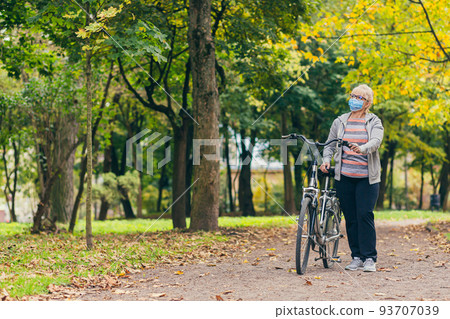 Senior beautiful blonde woman rides a bike in the park, in the woods, in nature in a medical mask during quarantine, leads a healthy active lifestyle Senior beautiful blonde woman rides a bike in the park, in the woods, in nature in a medical mask during quarantine, leads a healthy active lifestyle 93707039
