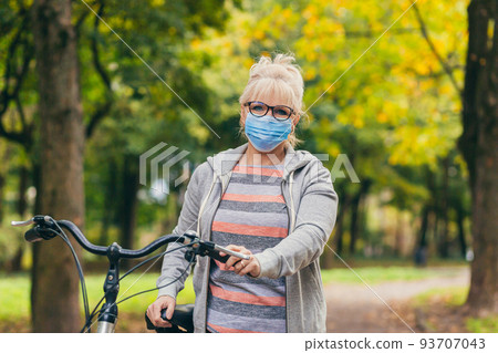 Senior beautiful blonde woman rides a bike in the park, in the woods, in nature in a medical mask during quarantine, leads a healthy active lifestyle Senior beautiful blonde woman rides a bike in the park, in the woods, in nature in a medical mask during quarantine, leads a healthy active lifestyle 93707043