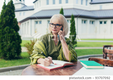 senior beautiful business woman sitting at a table outside, working online, using laptop, talking on the phone, writing in a notebook, working with documents 93707092