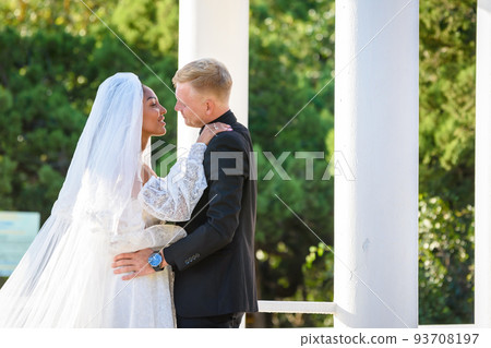 Mixed-racial newlyweds on a walk hugging and lovingly look at each other 93708197