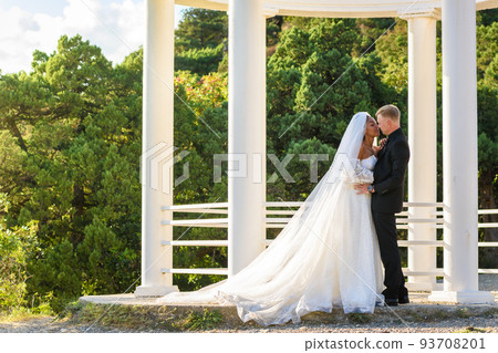 Portrait of mixed-racial newlyweds against the background of a gazebo with round columns, a girl in a lush white dress 93708201