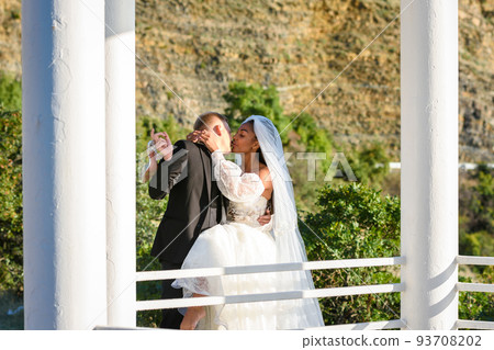 Young beautiful interracial newlyweds are kissing in the gazebo, against the backdrop of sunny mountains Young beautiful interracial newlyweds are kissing in the gazebo, against the backdrop of sunny mountains 93708202
