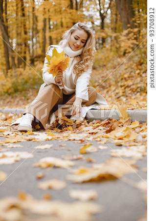 Young woman sitting on a curb in autumn forest. Blonde woman holding a yellow leaves. Girl wearing beige coat and brown trousers. Young woman sitting on a curb in autumn forest. Blonde woman holding a yellow leaves. Girl wearing beige coat and brown trousers. 93708912